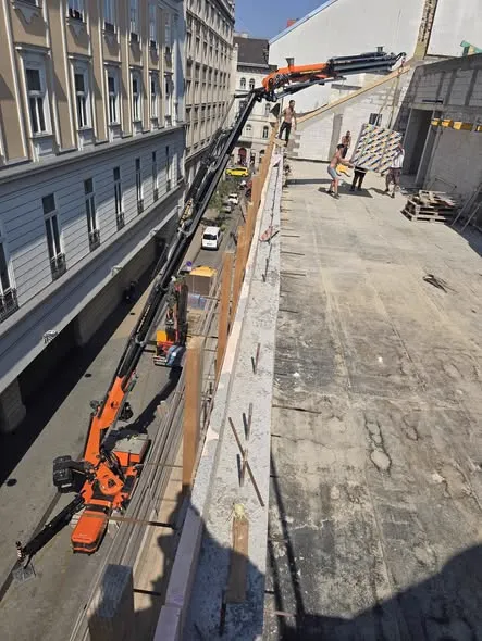 Frissen betonozott tetőszinti födém betonacél-kiállásokkal, mellette az utca fölé nyúló betonpumpa gémje, szemben klasszikus bérházhomlokzat